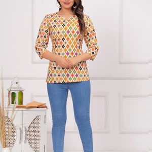 K&ouml;nnte beinhalten: Eine Frau tr&auml;gt ein wei&szlig;es Oberteil mit einem farbenfrohen geometrischen Muster und Dreiviertel&auml;rmeln, kombiniert mit hellblauen Skinny-Jeans. Das Oberteil hat einen Rundhalsausschnitt. Das Outfit wird durch transparente Absatzsandalen erg&auml;nzt.