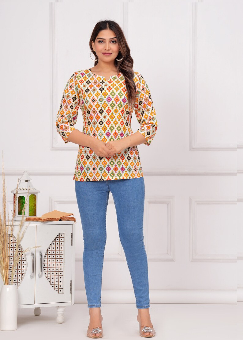 K&ouml;nnte beinhalten: Eine Frau tr&auml;gt ein wei&szlig;es Oberteil mit einem farbenfrohen geometrischen Muster und Dreiviertel&auml;rmeln, kombiniert mit hellblauen Skinny-Jeans. Das Oberteil hat einen Rundhalsausschnitt. Das Outfit wird durch transparente Absatzsandalen erg&auml;nzt.