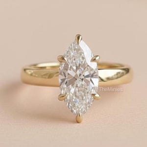 May include: A marquise-cut diamond engagement ring with a gold band. The diamond is set with four prongs and sparkles brightly. The ring is displayed on a light pink surface.