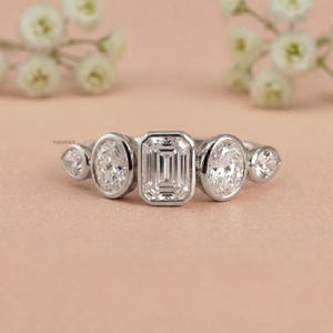 Peut inclure: Une bague en argent ornée d'une pierre centrale de taille émeraude, flanquée de deux pierres de taille ovale et de deux pierres plus petites. La bague est posée sur un fond rose clair avec des fleurs blanches.