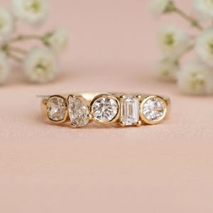 May include: A gold ring featuring five clear gemstones of varying shapes: oval, pear, round, emerald cut, and round. The ring is set against a soft pink background, with white flowers in the background.
