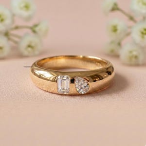 Könnte beinhalten: Ein Goldring mit zwei eingelassenen, klaren Edelsteinen, einem im Smaragdschliff und einem im Tropfenschliff, die nebeneinander angeordnet sind. Das Metall hat eine polierte, glatte Oberfläche und das Wort THEMINI ist klein schwarz innen eingraviert.