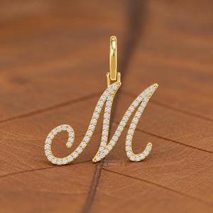 May include: A gold-toned letter M pendant featuring a cursive script design encrusted with small, clear, sparkling stones. The pendant includes a vertical bail at the top for attachment to a chain. The text THE MINIES is visible.