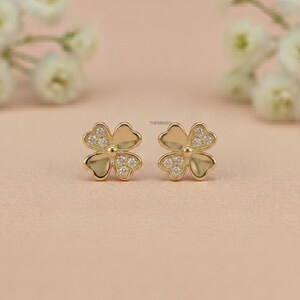 May include: A pair of gold-colored clover-shaped stud earrings. Each earring features four heart-shaped petals with small, clear gemstones in the center. The earrings are set against a soft pink background.