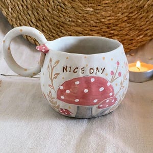Op de afbeelding: Handbeschilderde keramische mok met een grillig paddenstoelontwerp. De mok heeft een grote rode paddenstoel met witte stippen, de woorden "NICE DAY" en een gebogen handvat met gouden stippen. De mok is gebroken wit met bloemaccenten.