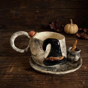 May include: A ceramic mug and saucer set with a mottled grey finish. The mug features a sculpted black witch hat on the side, a round handle with a small orange pumpkin accent, and the word HALLOWEEN embossed on the saucer rim with a small cauldron.