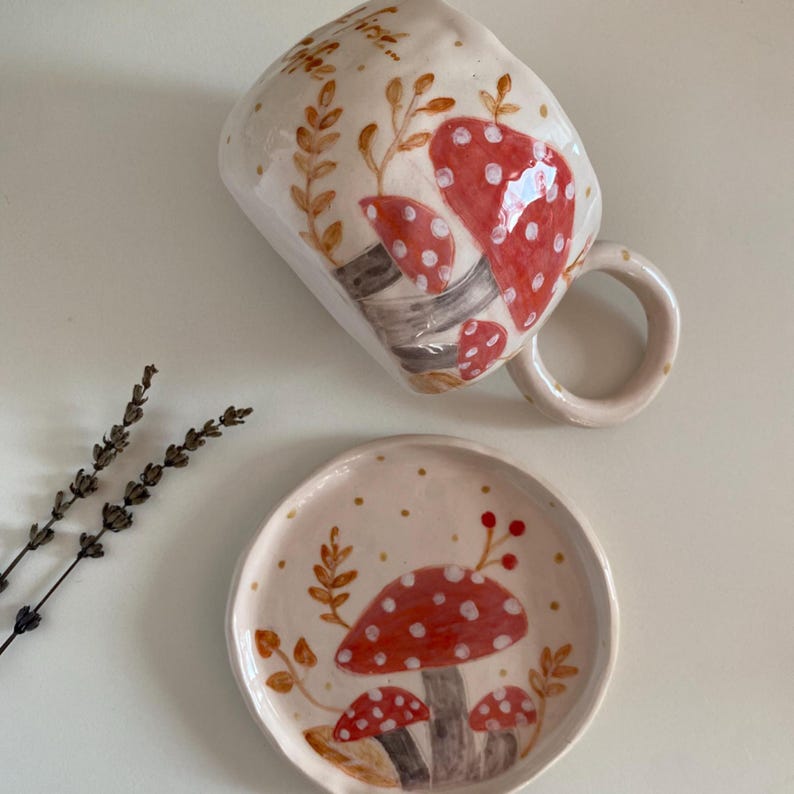 Op de afbeelding: Een handgemaakte keramische mok en schotel set. De mok en schotel zijn gebroken wit met een grillig ontwerp van rode paddenstoelen met witte stippen. De paddenstoelen hebben grijze stelen en zijn omgeven door bruine bladeren en gouden stippen.