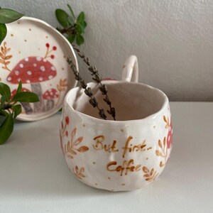 Op de afbeelding: Cr&egrave;mekleurige keramische mok met een handvat, met een handgeschilderd ontwerp van rode paddenstoelen en de tekst "But first... Coffee." Een bijpassend bord met een paddenstoelontwerp is zichtbaar. De mok bevat gedroogde lavendel.