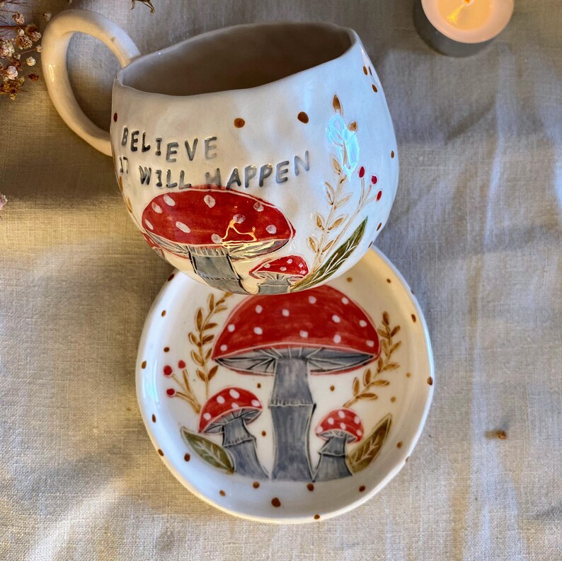 Puede incluir: Juego de taza y platillo de cer&aacute;mica blanca con un dise&ntilde;o caprichoso de setas. La taza presenta el texto "BELIEVE IT WILL HAPPEN" y est&aacute; adornada con setas rojas y blancas, puntos marrones y hojas verdes. El platillo refleja el tema de las setas.