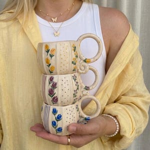 Op de afbeelding: Een stapel van drie crèmekleurige keramische mokken met verticaal geribbelde texturen en handgeschilderde bloemmotieven. Elke mok heeft een unieke kleur tulpen, waaronder geel, paars en blauw, naast fijne gouden of blauwe stippen.