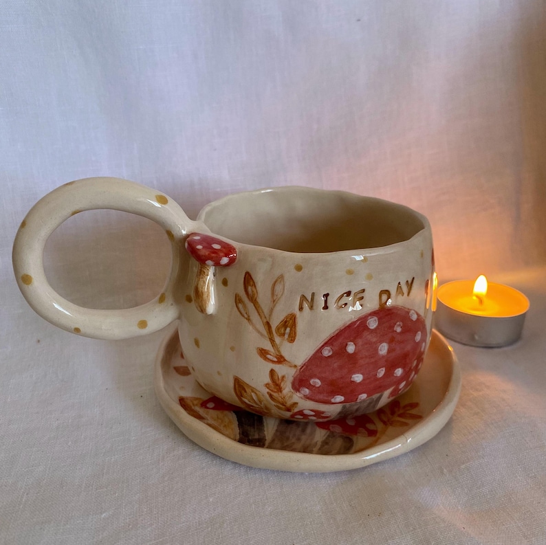 Op de afbeelding: Cr&egrave;mekleurige keramische mok en schotelset met een grillig paddenstoelontwerp. De mok heeft een grote handgreep met gouden stippen, een rood paddenstoelaccent en de tekst "NICE DAY". De schotel heeft een bijpassende paddenstoelillustratie.