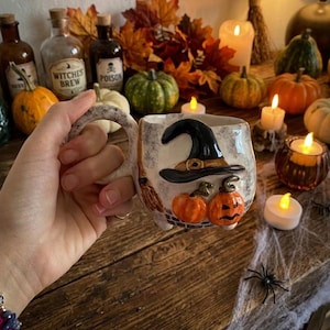 Può includere: Una tazza in ceramica con finitura bianco sporco maculata, caratterizzata da un cappello da strega nero in rilievo, una scopa e due zucche arancioni con volti intagliati. La tazza presenta un manico curvo e una superficie strutturata.
