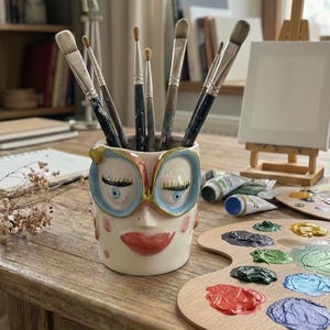 May include: A quirky ceramic owl-faced brush pot with blue-rimmed glasses, filled with paintbrushes. A wooden palette with various colours of paint and tubes of paint are nearby, along with a small easel and canvas, suggesting an artist's workspace.