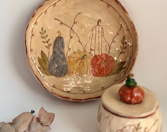 May include: A round ceramic plate with a cream-colored glaze featuring hand-painted illustrations of three pumpkins in blue, yellow, and orange, accompanied by a matching lidded ceramic jar topped with a small sculpted pumpkin finial.