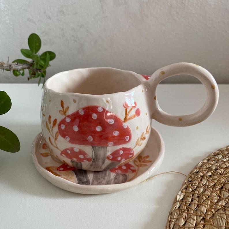 Op de afbeelding: Een keramische kop en schotel set. De kop en schotel zijn gebroken wit met een handgeschilderd ontwerp van rode paddenstoelen met witte stippen. De kop heeft een grote, afgeronde oor. De set is versierd met kleine gouden stippen en bruine bladeren.