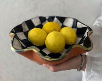 May include: A decorative ceramic bowl with a black and white checkered pattern interior and a terracotta-colored exterior. The bowl holds four bright yellow lemons. The rim is trimmed with gold paint, adding a touch of elegance to the design.