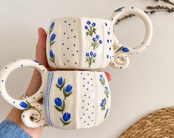 May include: Two cream-colored ceramic mugs with vertical ridges, each featuring hand-painted blue floral patterns, polka dots, and small hearts. The mugs have large, circular handles with a decorative swirl at the base and painted blue tulips.