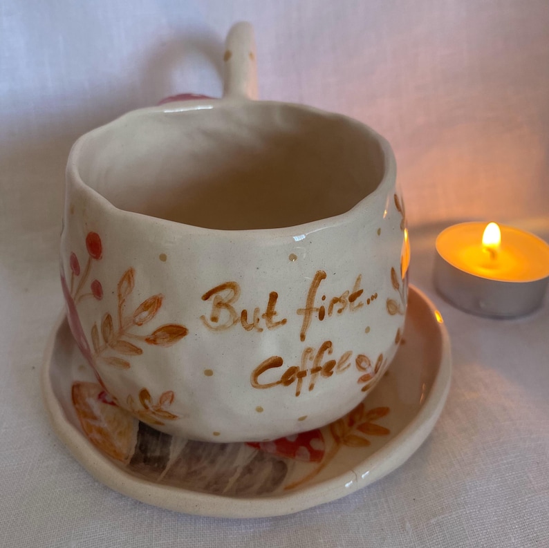 Op de afbeelding: Cr&egrave;mekleurige keramische koffiekop en schotel set. De beker heeft een handvat en is versierd met bruine bloemmotieven en de tekst "But first... Coffee". Naast de beker staat een brandende waxinelichtkaars.