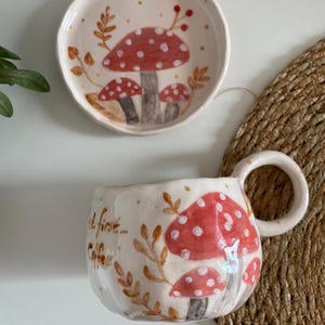 Op de afbeelding: Een keramische mok en kleine bordenset. De cr&egrave;mekleurige stukken zijn versierd met rode en witte paddenstoelillustraties en bruine bladaccenten. De mok heeft een grote handgreep en de woorden "At first... Coffee" staan op de zijkant.