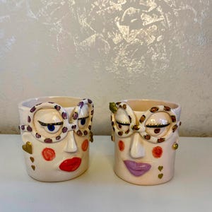 May include: Two ceramic mugs with painted faces, each with a pair of glasses and a unique expression. One mug has pink lips and red cheeks, while the other has purple lips and orange cheeks.