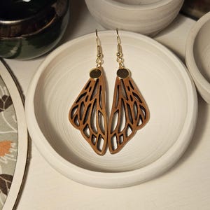Puede incluir: Pendientes de madera con diseño de alas de mariposa. Los pendientes tienen ganchos dorados y pequeños discos dorados. Las alas son de color marrón claro con un patrón recortado.