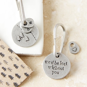 Op de afbeelding: Zilveren sleutelhanger met een bedel die "We'd be lost without you" zegt en een bedel met een klein boomontwerp. De sleutelhanger heeft ook een klein hartbedel.