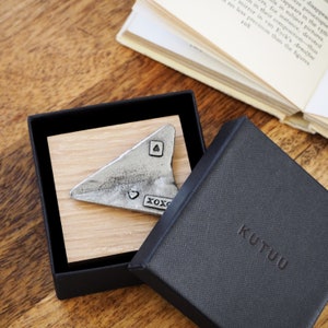 May include: A small silver airplane charm with the words "XOXO" and a heart engraved on it. The charm is displayed in a black box with the word "KUTUU" printed on the bottom.
