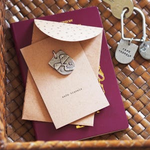 May include: A silver-toned metal charm featuring an embossed mountain and river landscape, attached to a tan card printed with the text safe travels. The card is tucked into a matching tan envelope, resting on a burgundy passport and a metal keychain.