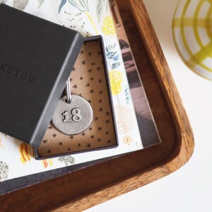 May include: A small, silver, round keychain with the number 18 on it. The keychain is in a brown box with a white and black patterned design. The box is open and the keychain is visible. The box is sitting on a wooden surface.