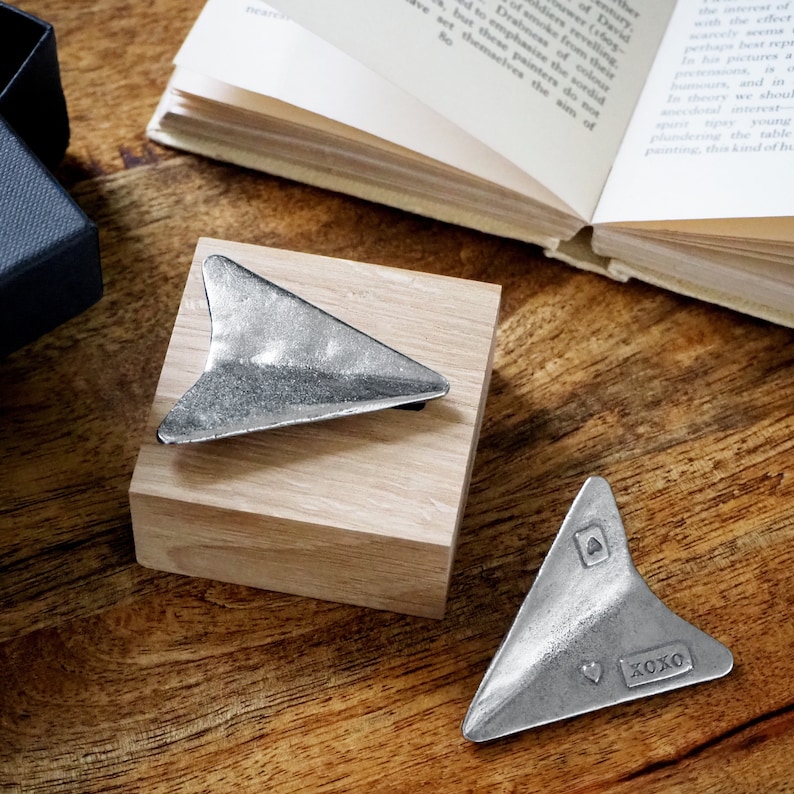May include: Two silver paper airplane shaped trinkets. One is on a wooden block and the other is on a wooden table. The smaller airplane has the word "XOXO" engraved on it.