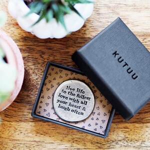 May include: A silver round token with the words "live life to the fullest love with all your heart & laugh often" engraved on it. The token is in a small black box with a white heart pattern and the word "KUTUU" printed on the box.