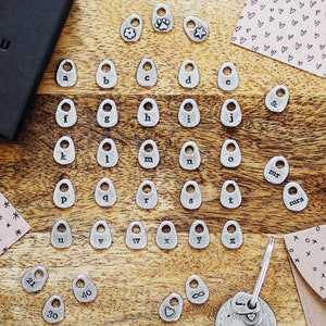 Könnte beinhalten: Eine Sammlung kleiner, silberfarbener Anhänger und Anhänger, darunter Buchstaben, Zahlen und Symbole wie ein Pfotenabdruck, ein Stern und das Unendlichkeitszeichen. Die Anhänger sind auf einer Holzoberfläche angeordnet.