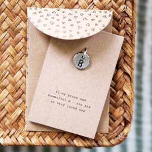 Könnte beinhalten: Eine braune Karte mit dem Text "to my brave and beautiful B - you are so very loved xxx" und einem silbernen Anhänger mit dem Buchstaben "B".