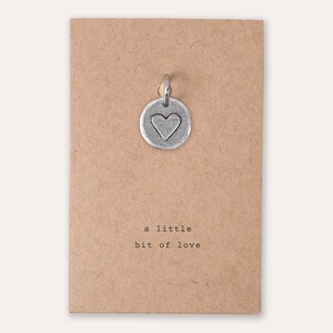 Peut inclure: Un charme argent&eacute; en forme de c&oelig;ur sur une carte brune avec l'inscription "a little bit of love".