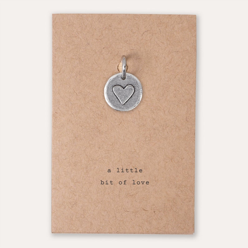 Peut inclure: Un charme argent&eacute; en forme de c&oelig;ur sur une carte brune avec l'inscription "a little bit of love".
