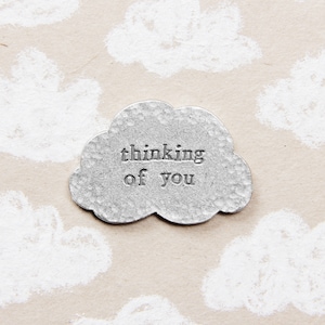 Pode incluir: Um amuleto de nuvem de metal prateado com o texto "thinking of you" em relevo. O amuleto está sobre um fundo marrom claro com formas de nuvens brancas.