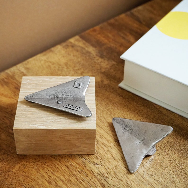 May include: Two silver metal arrow-shaped trinkets with the word "XOXO" engraved on one of them. The trinkets are sitting on a wooden block.