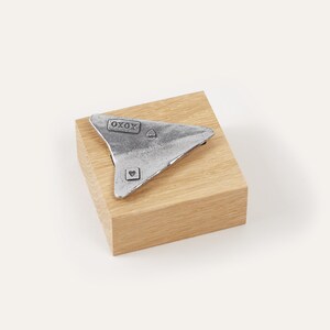 May include: A silver paper airplane with the word "XOXO" and a heart on it, sitting on a wooden block.