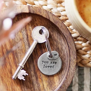 May include: A silver key with a round silver charm attached to it. The charm has the words "you are loved" engraved on it. The key and charm are sitting on a wooden surface.
