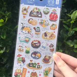 May include: A sheet of stickers featuring adorable cartoon cats enjoying various Japanese sweets and desserts. The stickers are colourful and feature a variety of designs, including shaved ice, mochi, dango, and green tea ice cream. The stickers are perfect for adding a touch of whimsy to your planner, journal, or laptop.