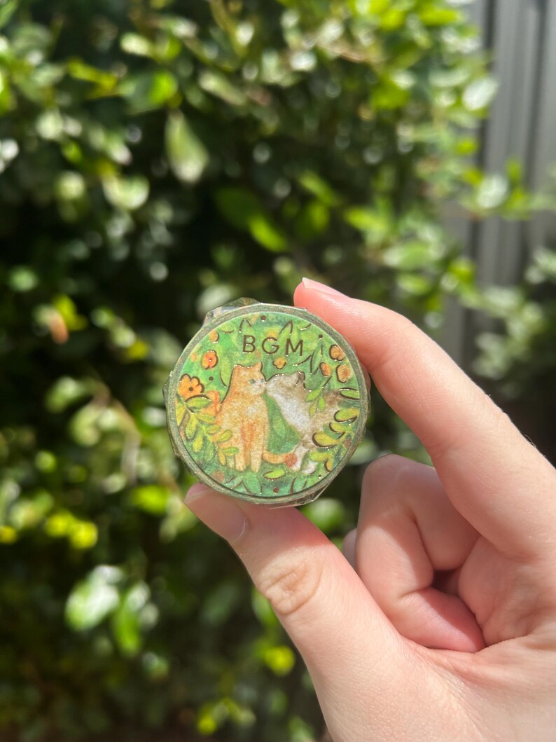 May include: A round, decorative container with a green and gold rim. The lid features a charming illustration of two cats, one orange and one gray, surrounded by flowers and foliage. The text "BGM" is also visible.