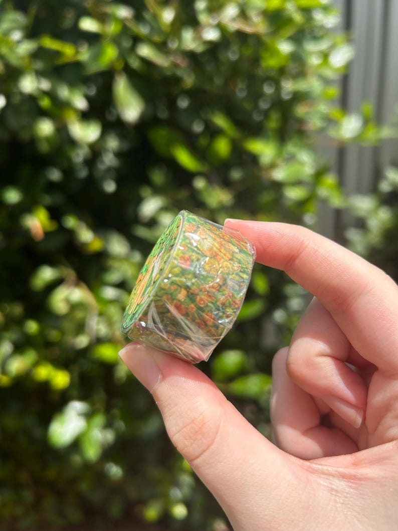 May include: A roll of decorative washi tape featuring a colorful floral design with green, orange, and yellow hues. The tape is wrapped in clear plastic, held by a hand against a backdrop of green foliage.