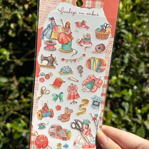 Può includere: Un foglio di adesivi con illustrazioni a tema cucito. Gli adesivi includono immagini di macchine da cucire, rocchetti di filo, forbici, vestiti e un orsacchiotto. Il testo "Fushigi na sekai" è scritto nella parte superiore del foglio.