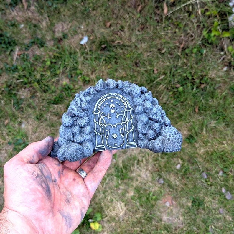 May include: A gray, stone-like sculpture of a stylized arched doorway with intricate gold-colored details. The doorway features a central emblem and is surrounded by textured rock formations. The sculpture is held in a hand against a backdrop of green grass.