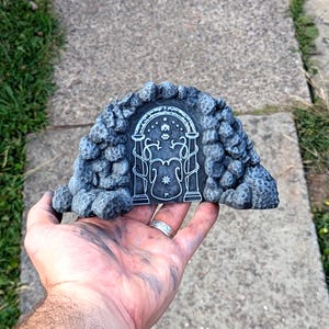 May include: A gray, arched doorway sculpture with a detailed stone texture. The door features a white, intricate design with floral and star motifs. The sculpture is held in a hand, showcasing its size and craftsmanship.