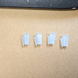 May include: Four white plastic pieces, each shaped like a small, open-ended box with a pointed top. The pieces are arranged in a row on a brown surface.