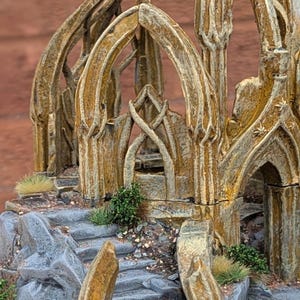 Peut inclure: Une miniature détaillée d'une structure fantastique avec des portes arquées et des marches en pierre. Le modèle présente un aspect vieilli et texturé avec des nuances de beige, de gris et de vert, suggérant un cadre ancien et envahi par la végétation.