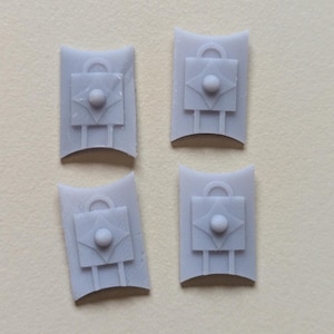 May include: Four small, gray, shield-shaped objects. Each has a central square design with a diamond and a sphere, a handle at the top, and two vertical lines at the bottom. The objects are arranged on a light background.