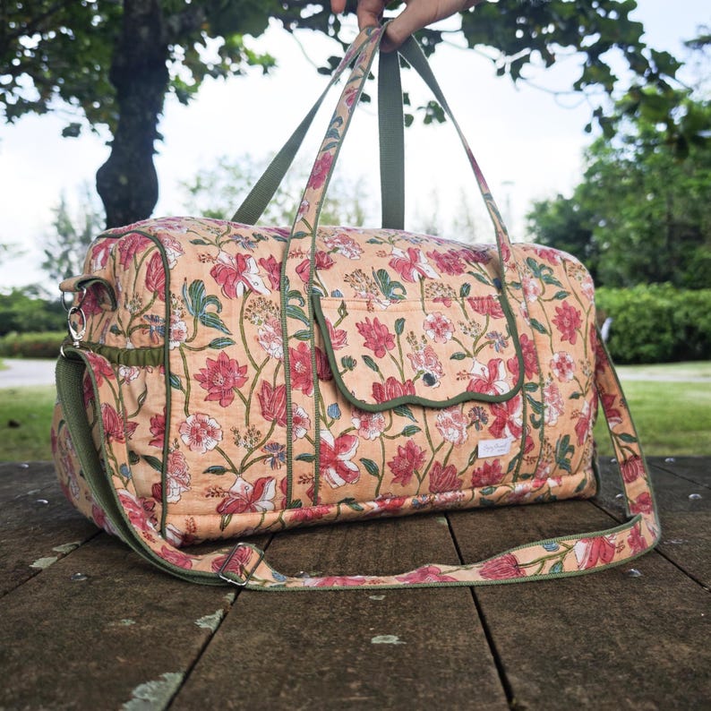 May include: A quilted duffel bag featuring a peach-colored fabric with a repeating floral pattern in shades of red, pink, and green. The bag includes olive green trim, two top handles, an adjustable shoulder strap, and a front pocket with a scalloped flap.