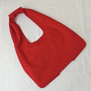 May include: A bright red tote bag with a single shoulder strap. The bag is made of a solid, smooth fabric and has a simple, minimalist design. The bag is empty and lies flat on a white surface.